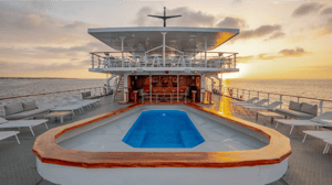 The upper deck of a cruise ship at sunset featuring a pool, lounge chairs, and bar area overlooking the ocean.